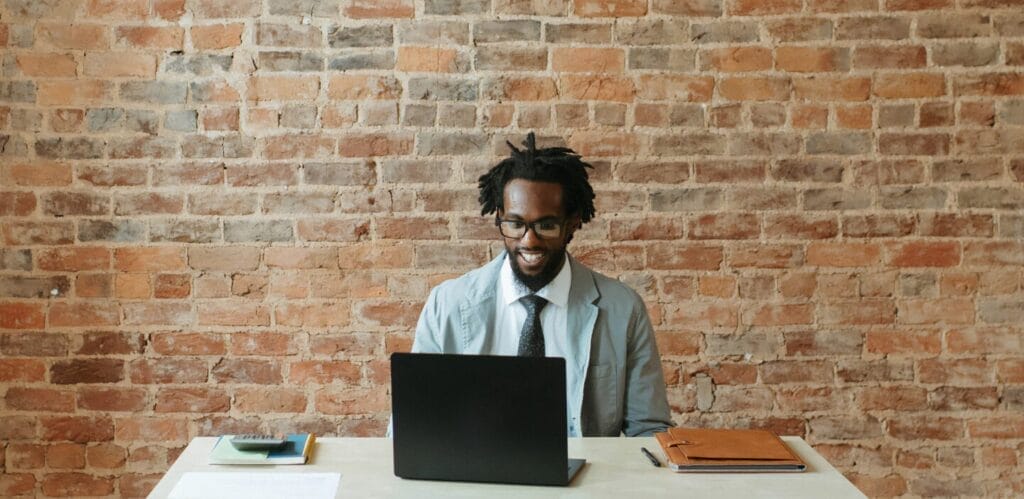 Man seated at a desk in front of a brick wall, and typing on his laptop